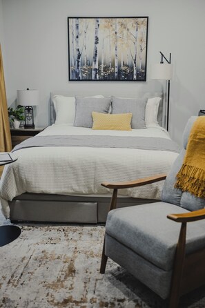 A bedroom with a bed, bedside tables, a lamp, a chair, a rug, and a painting on the wall. - Falkenberg Business Centre and Suites (Botwood)