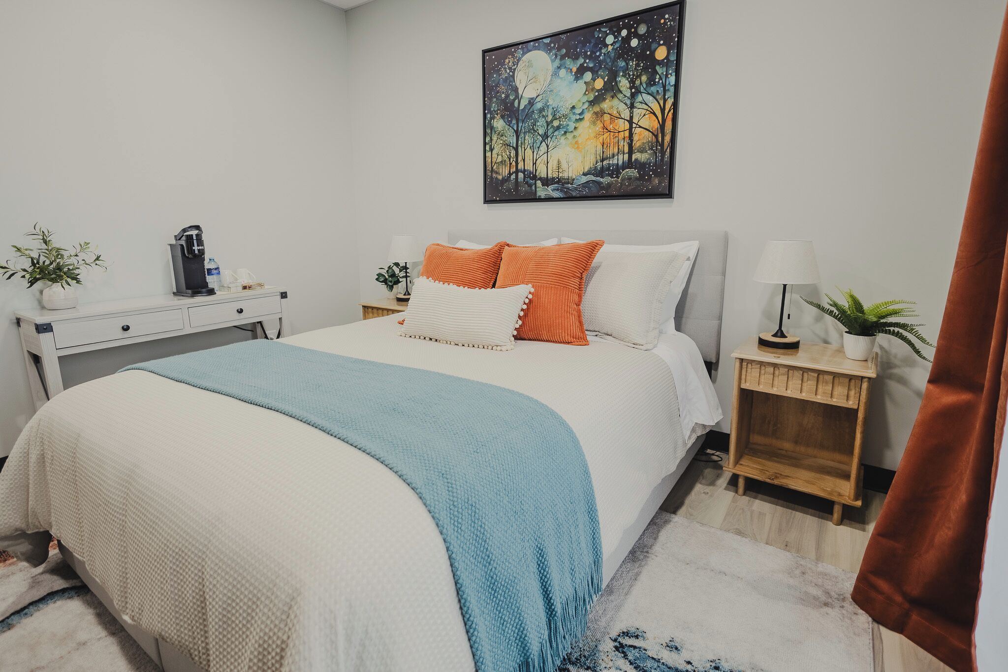 A neatly arranged bedroom with a bed, bedside tables, a coffee maker, and a painting on the wall.