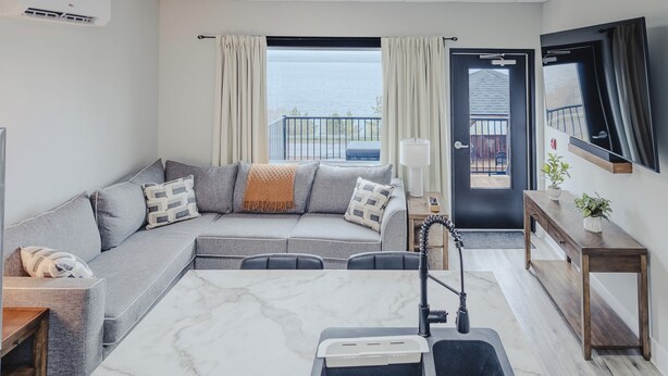 A modern living room with a grey sofa, a marble coffee table, a built-in kitchen with a sink, and a view of the sea through large windows. - Falkenberg Business Centre and Suites (Botwood)