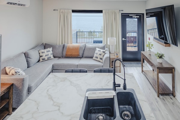 A modern living room with a grey sofa, a marble coffee table, a built-in kitchen with a sink, and a view of the sea through large windows.