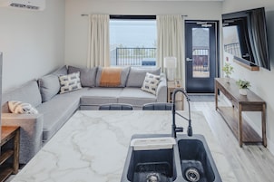 Un salón moderno con un sofá gris, una mesa de centro de mármol, una cocina integrada con un fregadero y vistas al mar a través de amplios ventanales.