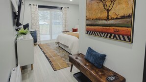 A hotel room with a bed, a bench, a TV, a painting, and a view of the outdoors. - Falkenberg Business Centre and Suites (Botwood)