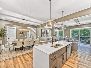 A modern kitchen with a central island, a dining area with a chandelier, and a living space with a sofa and large windows. - Modern Coastal Home with Private Pool and Free Golf Cart (Hilton Head Island)