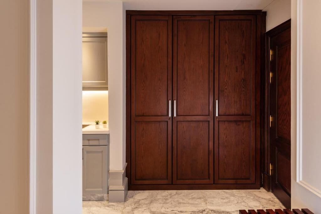 Un couloir avec une double porte en bois, un sol en marbre et un coin cuisine équipé de placards et d’un plan de travail.