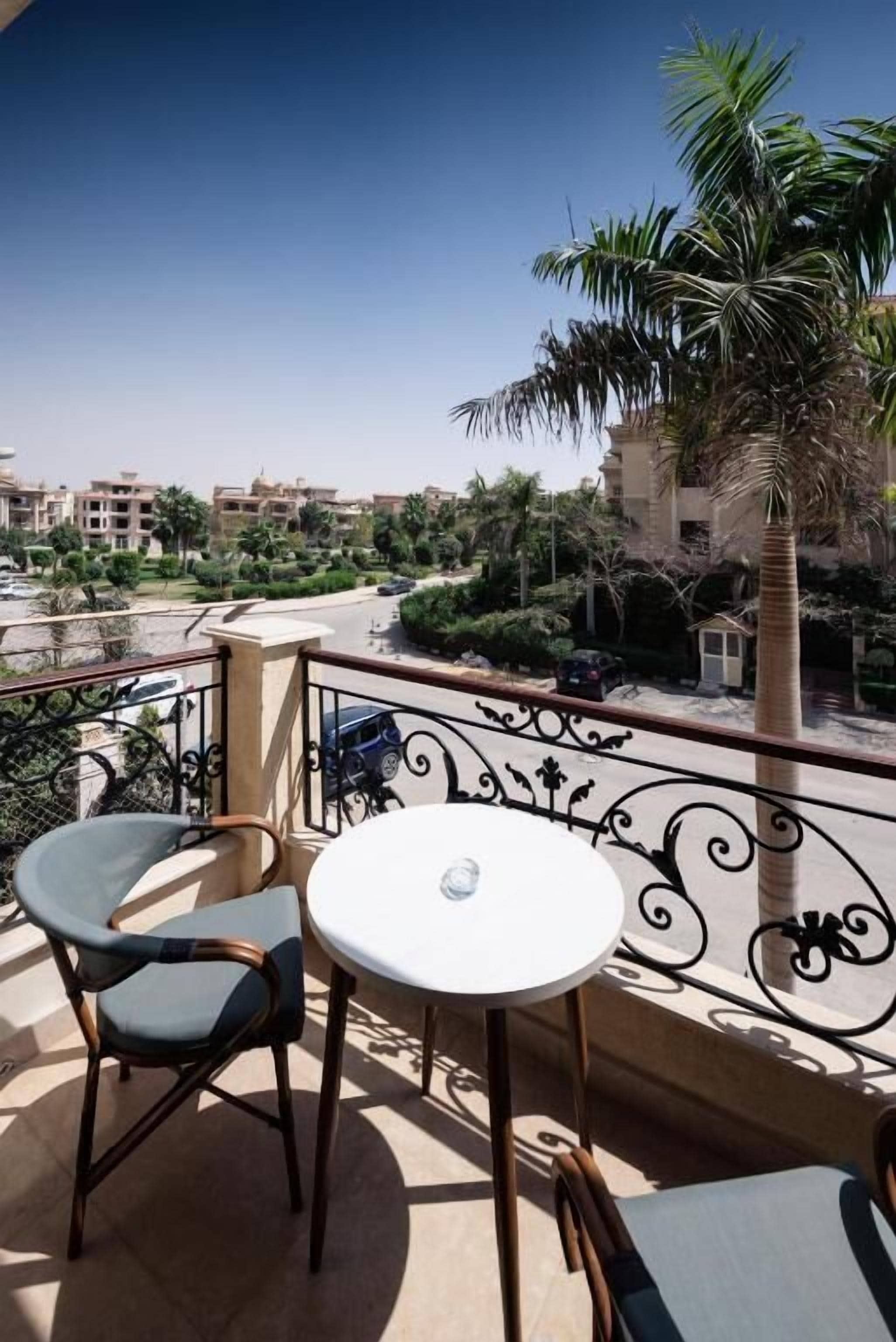 A balcony with two chairs and a table, overlooking a residential area with palm trees and buildings.