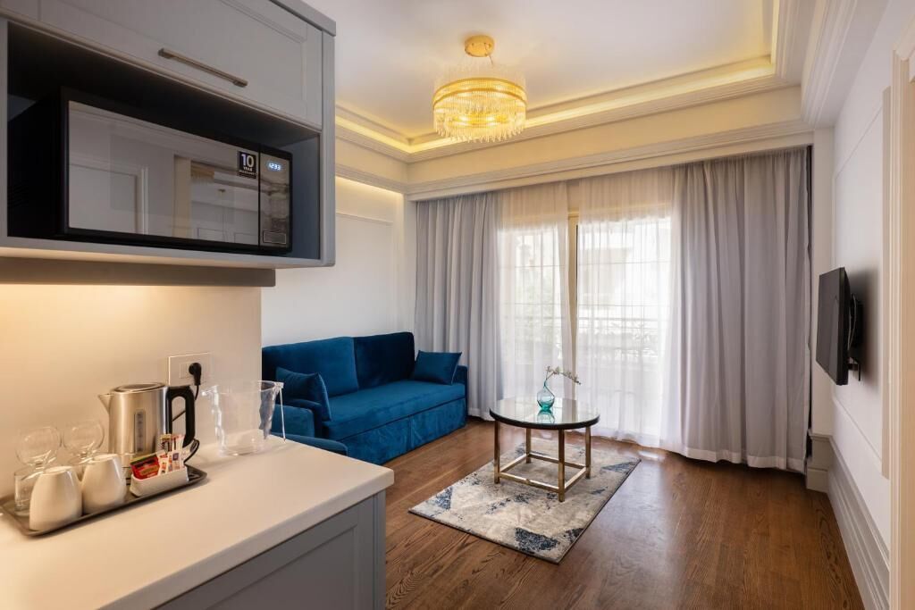 Una habitación de hotel moderna con kitchenette, un sofá azul, una pequeña mesa redonda y un amplio ventanal con cortinas.