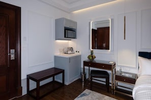 A hotel room with a wooden door, a desk with a mirror, a bench, a bed, and a small kitchenette area.