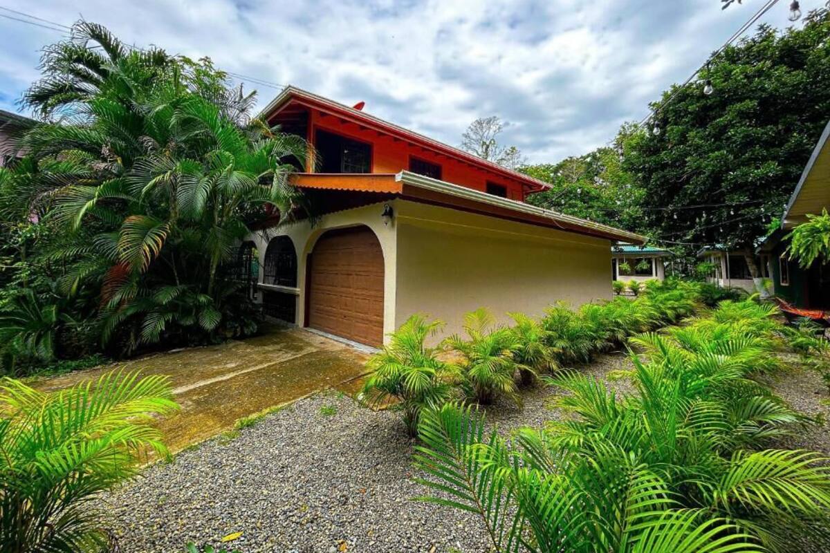 Una casa con techo rojo, puerta de garaje marrón y abundante vegetación alrededor.