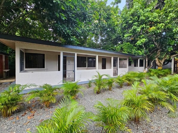 A white building with multiple windows and a covered walkway, surrounded by lush greenery and trees.