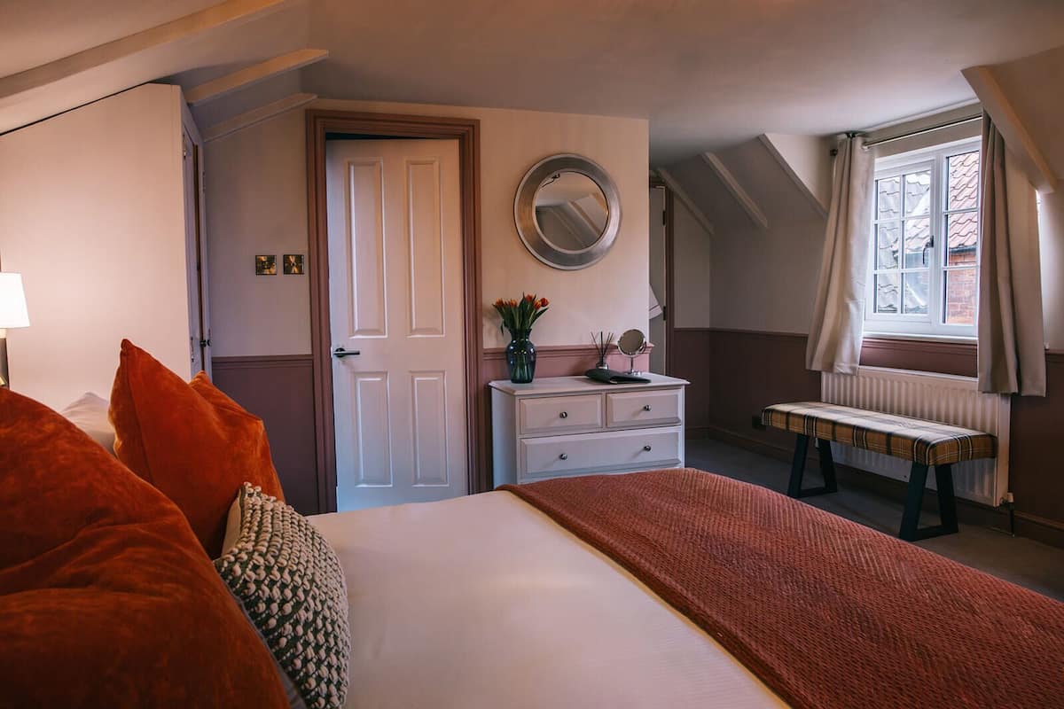 A bedroom with a bed, a dresser, a bench, a window with curtains, and a door.