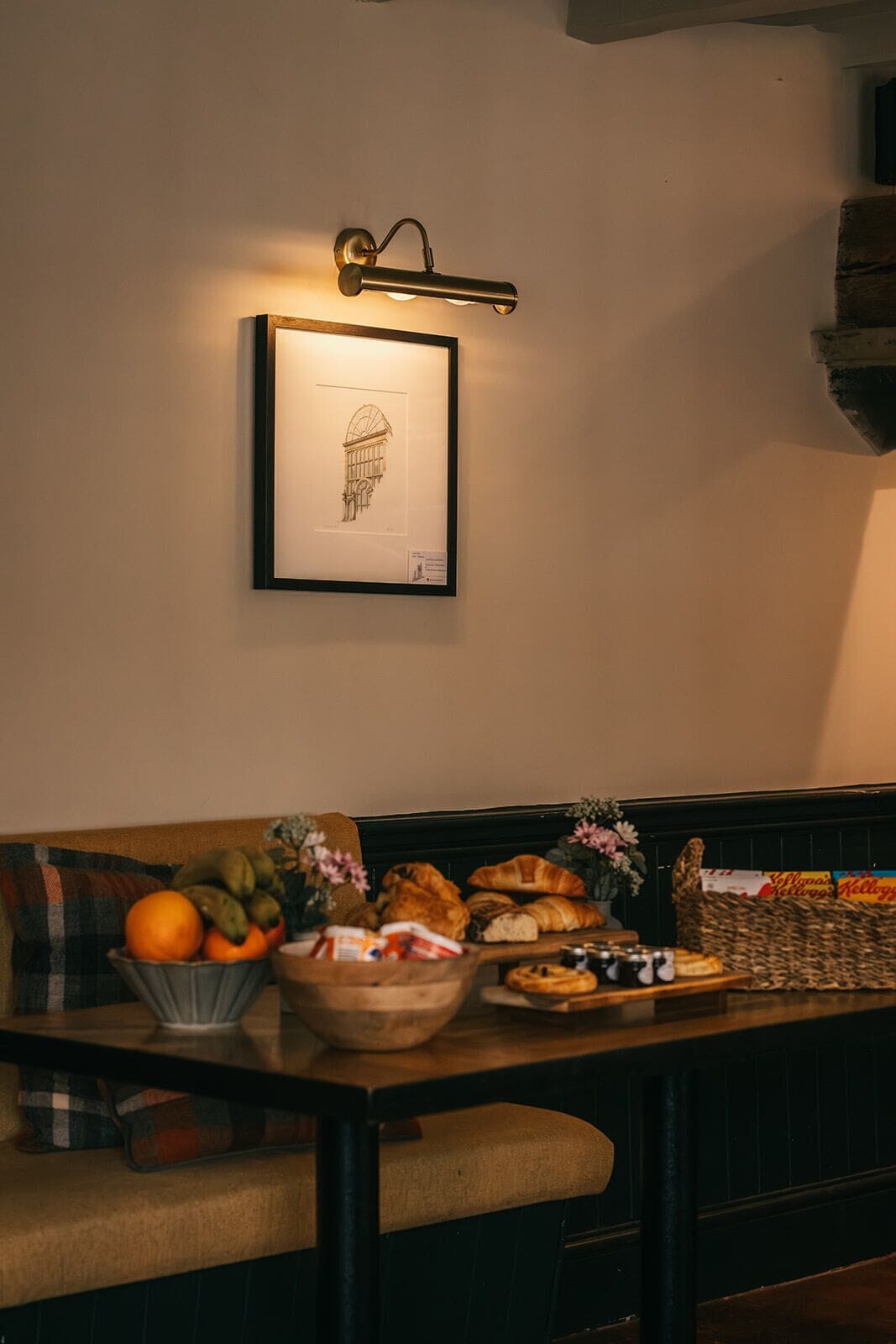 Un área de comedor con una mesa dispuesta para una comida, un dibujo arquitectónico enmarcado en la pared, y un banco con cojines.