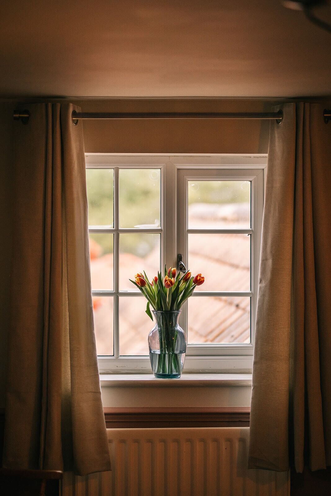 A window with a view of a wooden deck, curtains, and a vase with flowers.