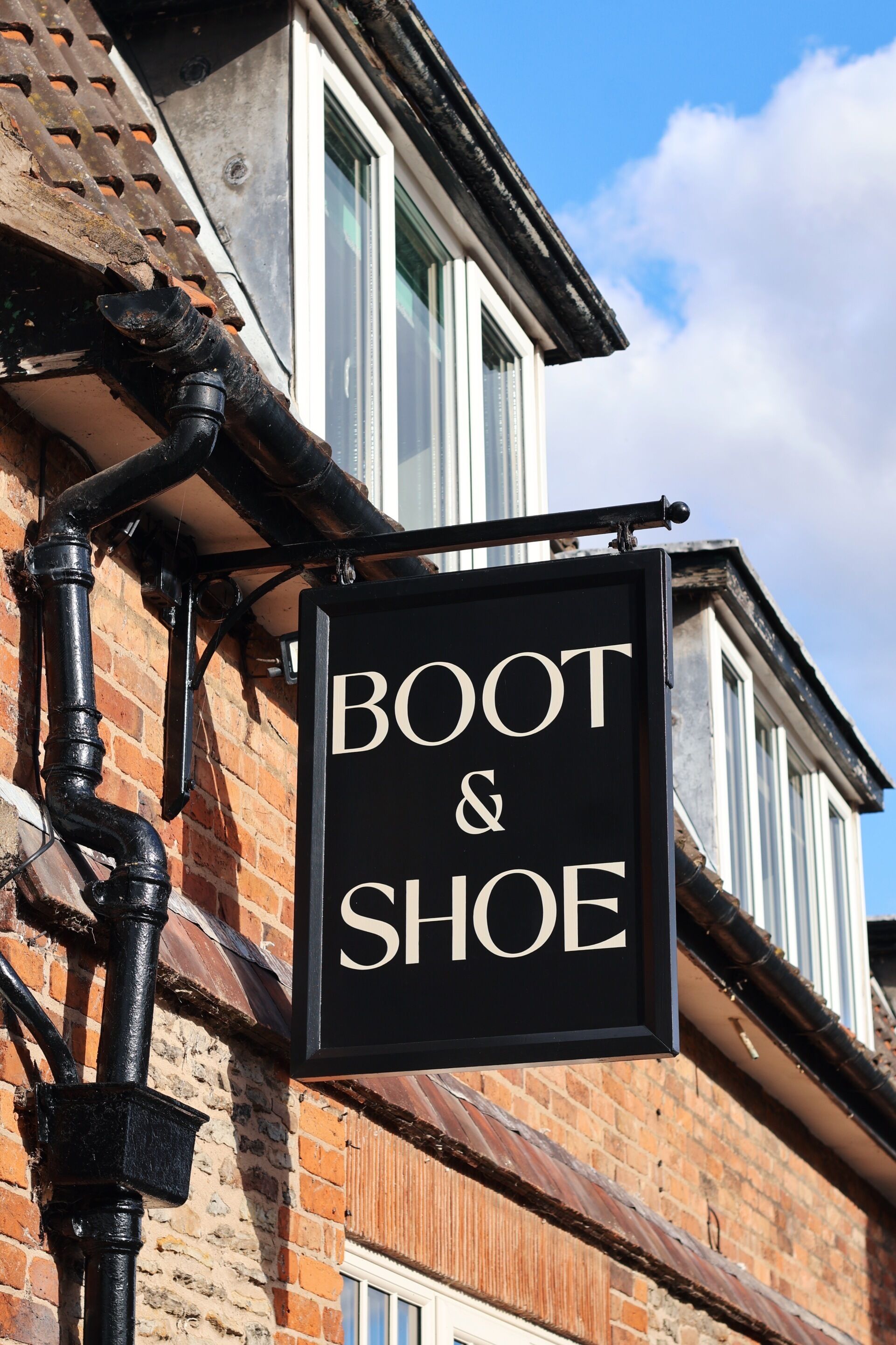 A black sign with 'BOOT & SHOE' written on it, attached to a brick building with a window and a metal gutter.