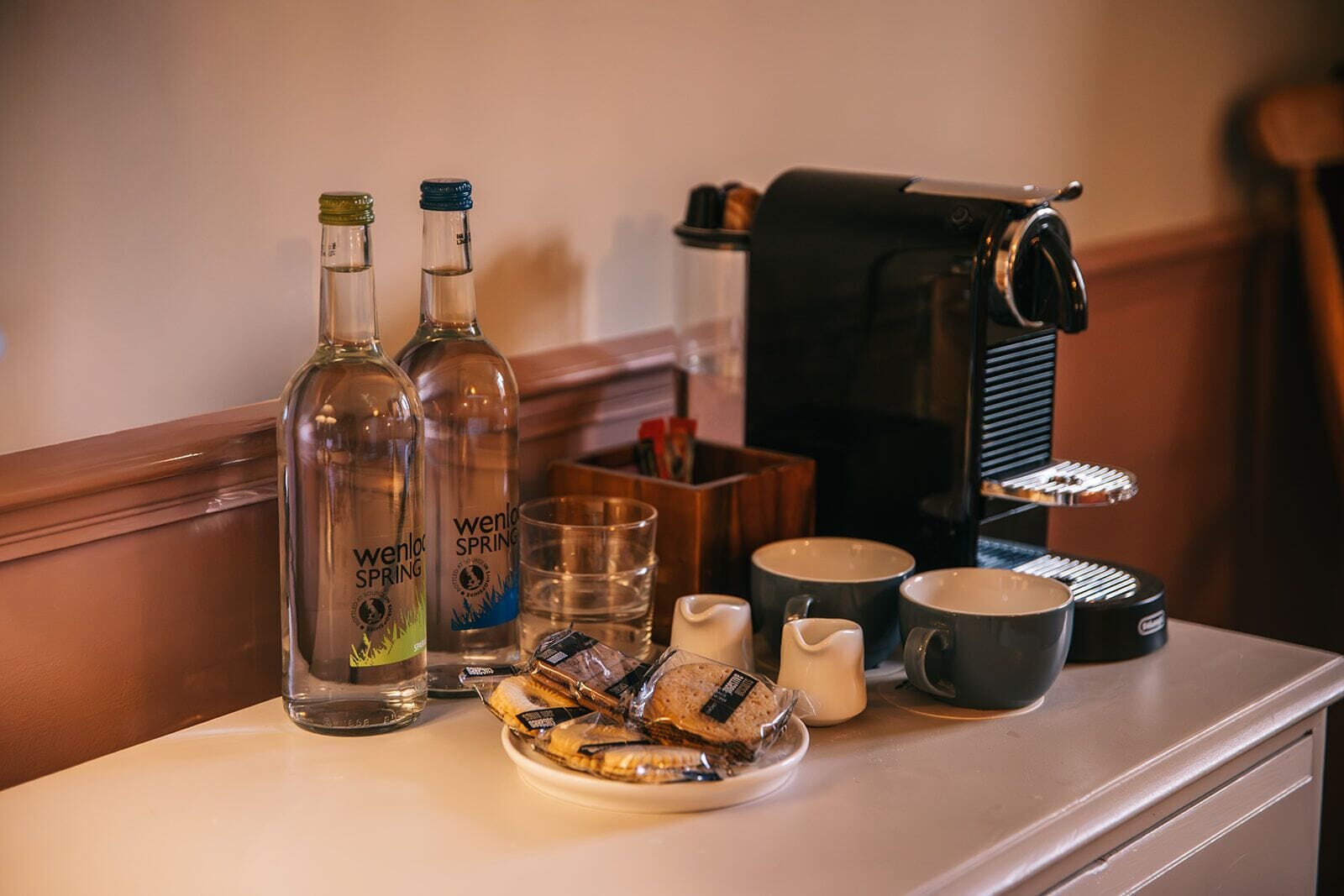 Una mesa con dos botellas etiquetadas 'Wenlock Spring', un vaso de agua, un plato de pasteles y una máquina de café.