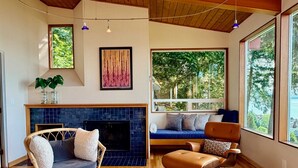 A cozy living room with a fireplace, a large window with a view of trees, and a patterned coffee table. - Luxury Bay Retreat Hot Tub Views (Bellingham)
