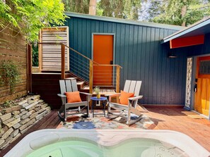 A backyard with a hot tub, two rocking chairs, a small table with a potted plant, and a wooden staircase leading to a door. - Luxury Bay Retreat Hot Tub Views (Bellingham)