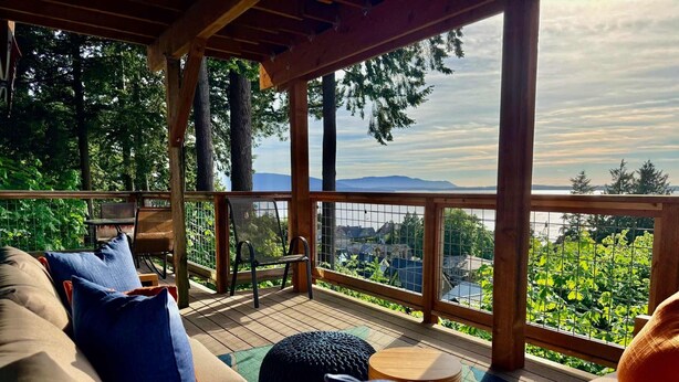 A covered deck with a sofa, chairs, and a table, overlooking a scenic view of trees and a body of water. - Luxury Bay Retreat Hot Tub Views (Bellingham)