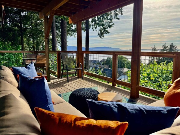 A covered deck with a sofa, chairs, and a table, overlooking a scenic view of trees and a body of water. - Luxury Bay Retreat Hot Tub Views (Bellingham)