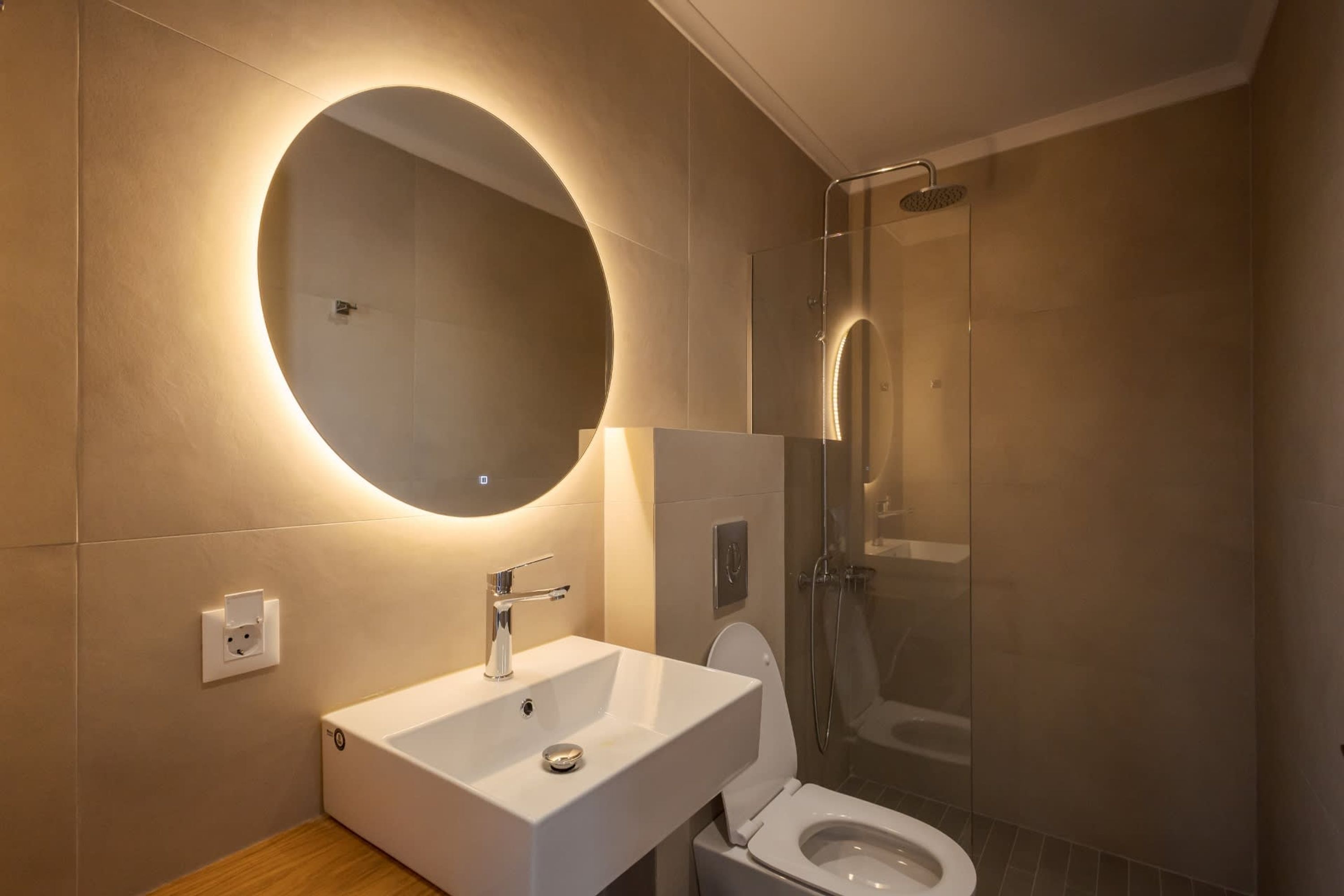 A modern bathroom with a round mirror, a white sink, a toilet, and a glass-enclosed shower.