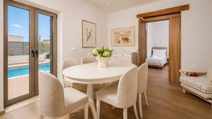 A modern interior with a round dining table, beige chairs, a white vase with flowers, a bed, and a view of a swimming pool. - Bon Filius - Luxury Private Villa w Pool in Chania (Apokoronas)