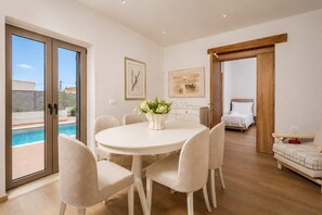 A modern interior with a round dining table, beige chairs, a white vase with flowers, a bed, and a view of a swimming pool. - Bon Filius - Luxury Private Villa w Pool in Chania (Apokoronas)