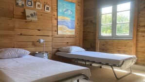 A room with two beds, a nightstand with a lamp, wooden walls, a painting, and a window with a view. - Holiday Home “Dormitory in Charente” with Private Terrace and Shared Garden (Jarnac)