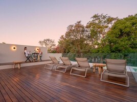 Terraza de madera con sillones y una mesa, rodeada de follaje y una piscina.