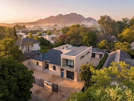 A modern residential area with houses featuring flat roofs and solar panels, surrounded by greenery and mountains in the background.