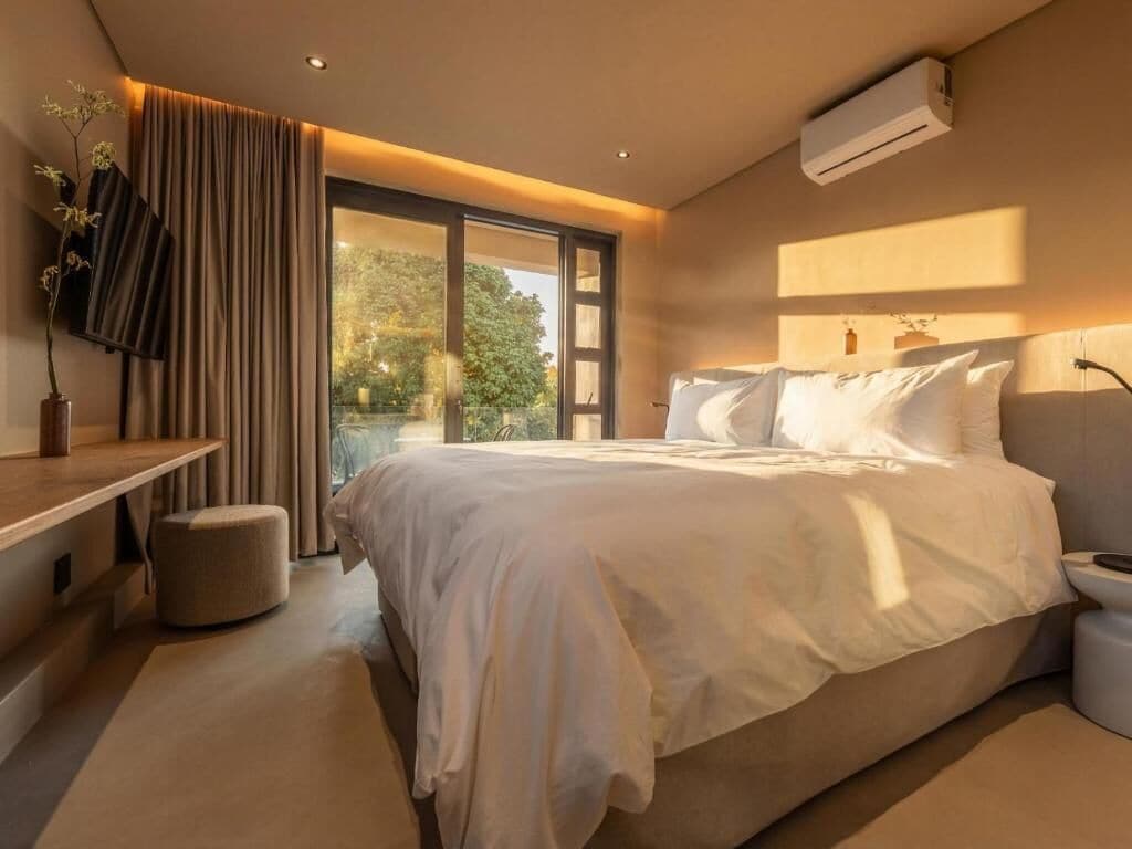 Une chambre à coucher moderne avec un grand lit, une table de chevet, un petit tabouret et une fenêtre avec des rideaux.