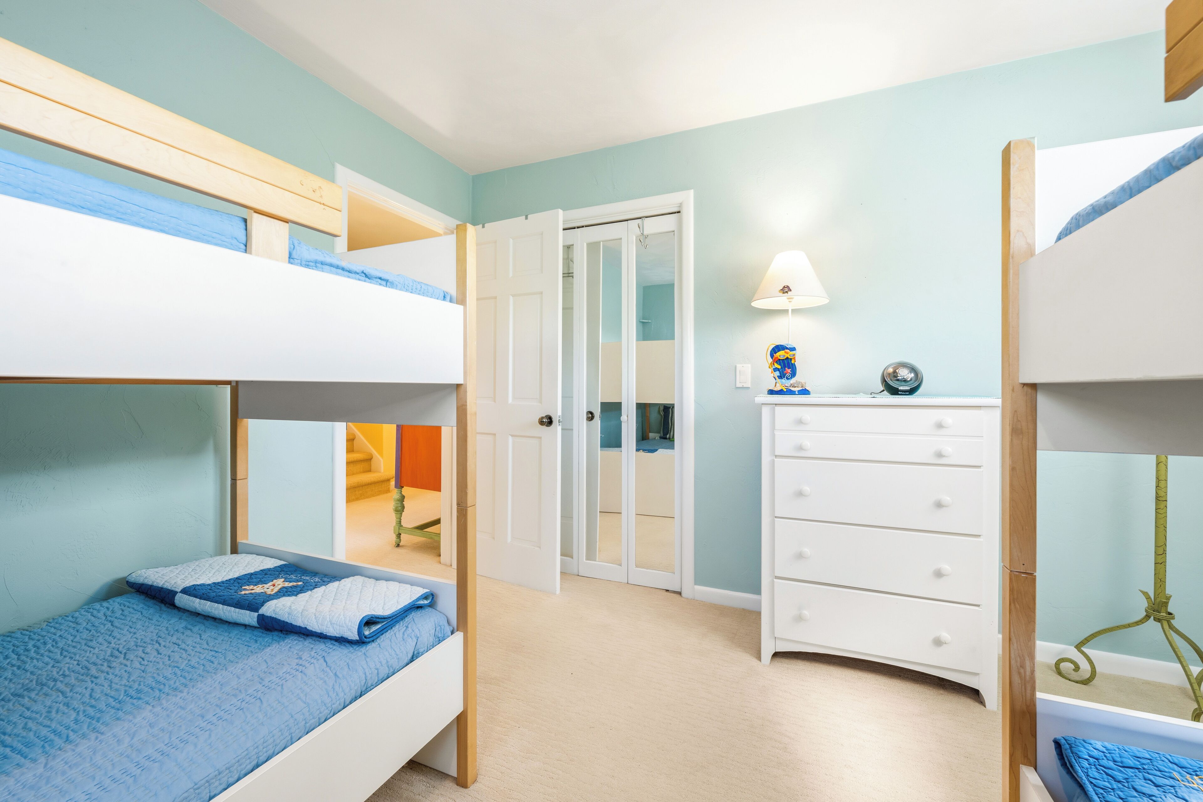 A bunk bed room with a white dresser, a door leading to another room, and a lamp on a side table.