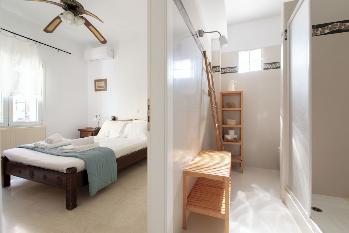 Ein Schlafzimmer mit Bett, Nachttisch, Ablagefläche, Deckenventilator, Fenster mit Vorhängen sowie ein Badezimmer mit Dusche, Holzregal und Handtuchhalter.