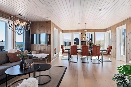 A spacious living area with a wooden ceiling, a fireplace, a dining area with chairs, and a large window offering a view.. Apartment for year-round holidays in Golsfjellet