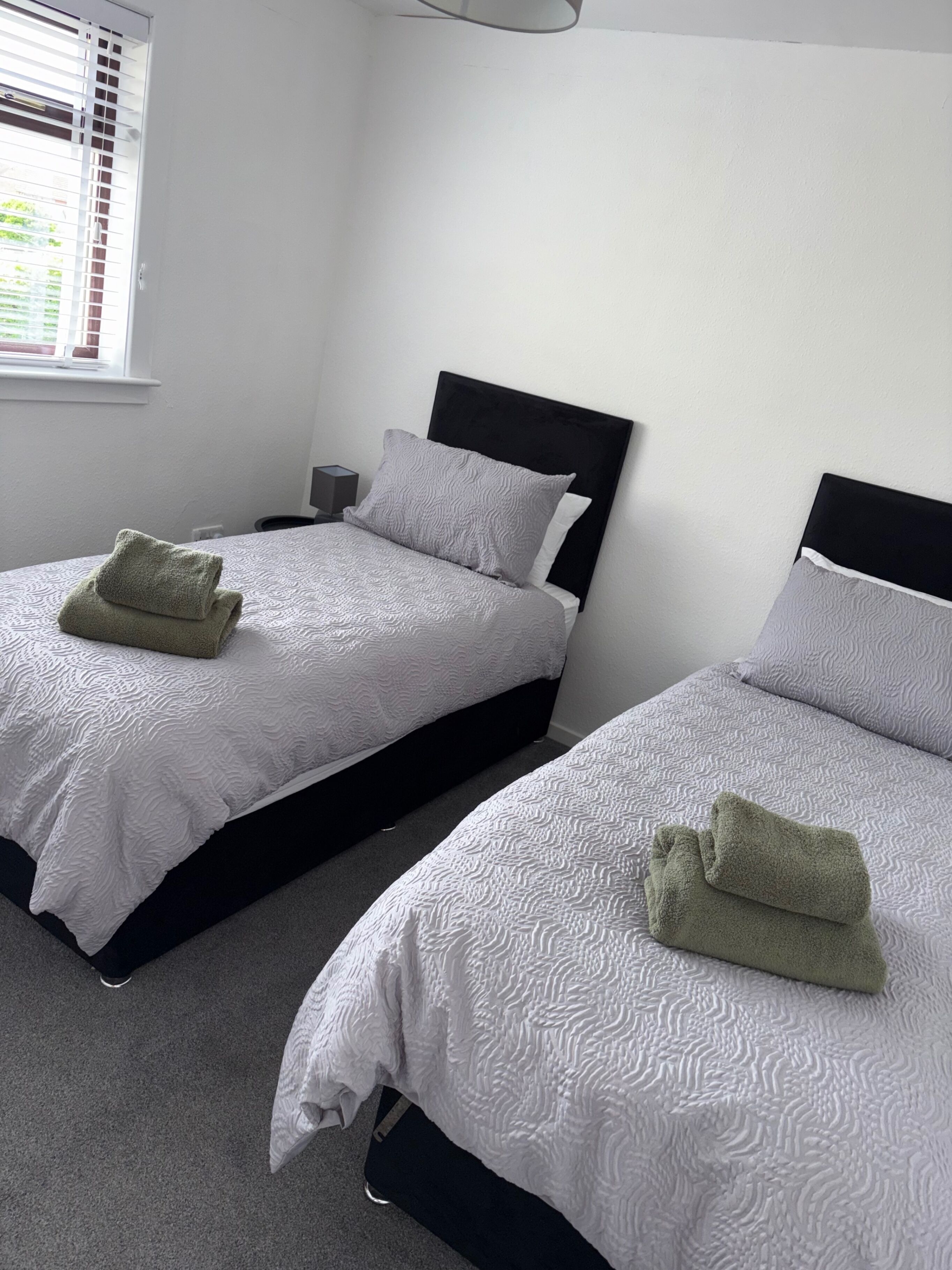 Two twin beds with white textured bedding and black headboards in a room with white walls and a window with blinds.