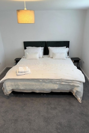A neatly made bed with white linens and pillows, a black headboard, a small bedside table with a lamp, and a grey carpeted floor.