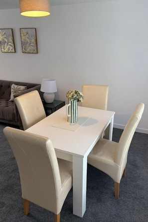 A dining area with a white table, beige chairs, a vase with flowers, a lamp, and a sofa in the background.