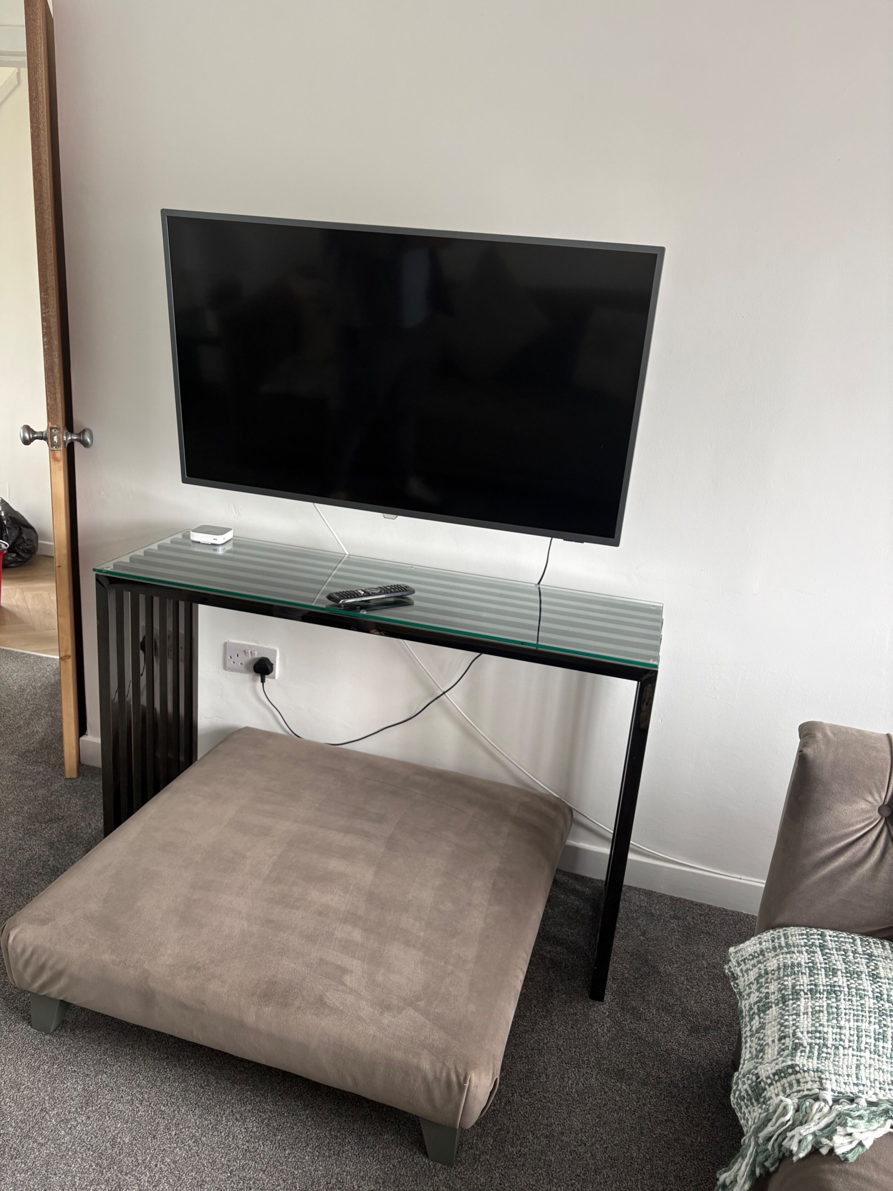 Een kamer met een wandgemonteerde flatscreen televisie, een bureau met glazen blad, een stopcontact, een voetenbankje en een groene en witte plaid.