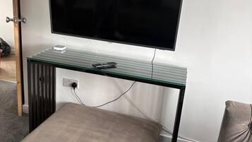 Een kamer met een wandgemonteerde flatscreen televisie, een bureau met glazen blad, een stopcontact, een voetenbankje en een groene en witte plaid.