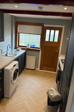 A compact kitchen with a built-in washing machine, a radiator, and a window with a view of a backyard. - Ayr 2-bed House, Sleeps 4 With Garden & Parking (Ayr)