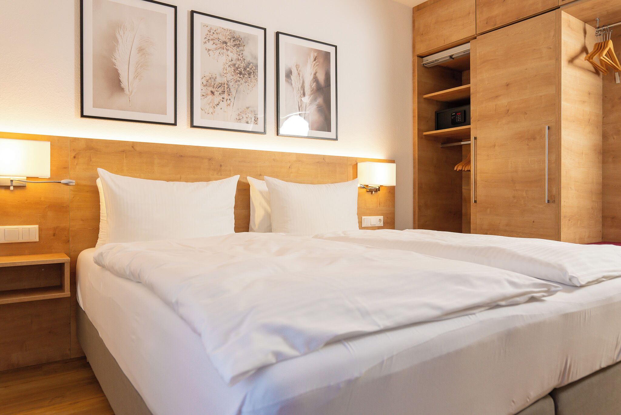 Ein Hotelzimmer mit einem Holzkopfteil, weißer Bettwäsche, einem Nachttisch mit Lampe und einem Kleiderschrank mit Kleidung.