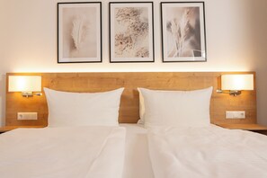 Cama doble con ropa de cama blanca, dos lámparas fijadas a la pared y tres cuadros con motivos florales.