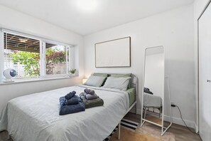 A bedroom with a bed, a window with blinds, a bedside table with a lamp, a wall-mounted mirror, and a radiator.