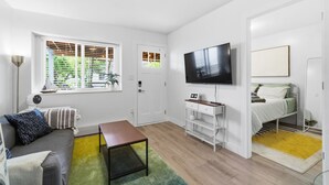 Un espacio habitable compacto con sofá, una mesita, televisor montado en la pared, cama con colchón y almohadas, y ventana con persianas.
