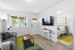 Un espacio habitable compacto con sofá, una mesita, televisor montado en la pared, cama con colchón y almohadas, y ventana con persianas.