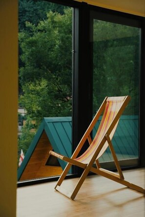 A wooden beach chair with a striped cushion, positioned in front of a large window with a view of green trees. - DRO Hotel (Kutaisi)
