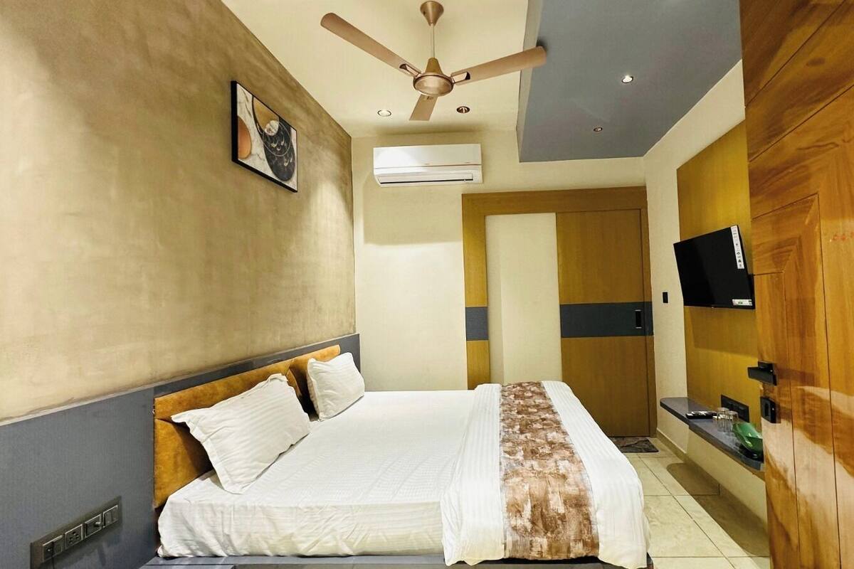 A hotel room with a bed, ceiling fan, air conditioning unit, wooden wardrobe, and a television.