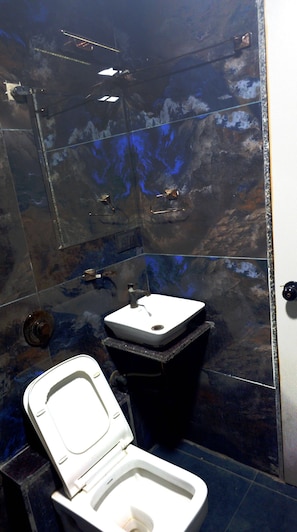 Un baño con inodoro blanco, un bote de basura negro y un lavamanos con espejo.