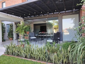 An outdoor patio with chairs and tables, surrounded by potted plants and a glass wall. - 5 Guest House Spacious: Quincho Swimming Pool Games (San Bernardino)
