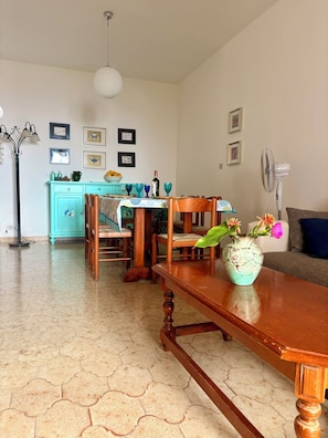 Un coin repas avec une table en bois, des chaises, un vase avec des fleurs, une cuisine en arrière-plan et un ventilateur sur la droite.