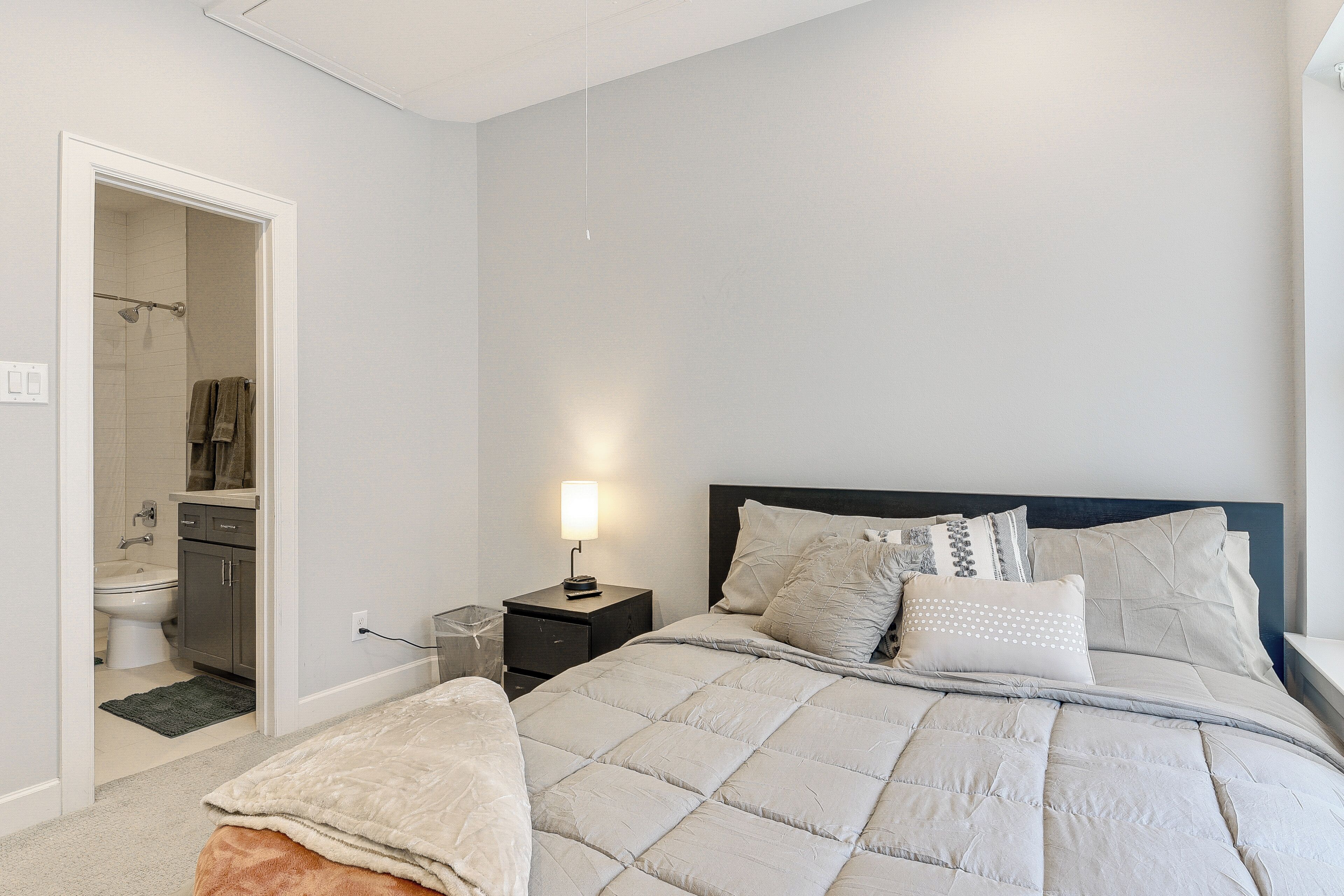 A bedroom with a bed, bedside table, lamp, and a view into the bathroom.