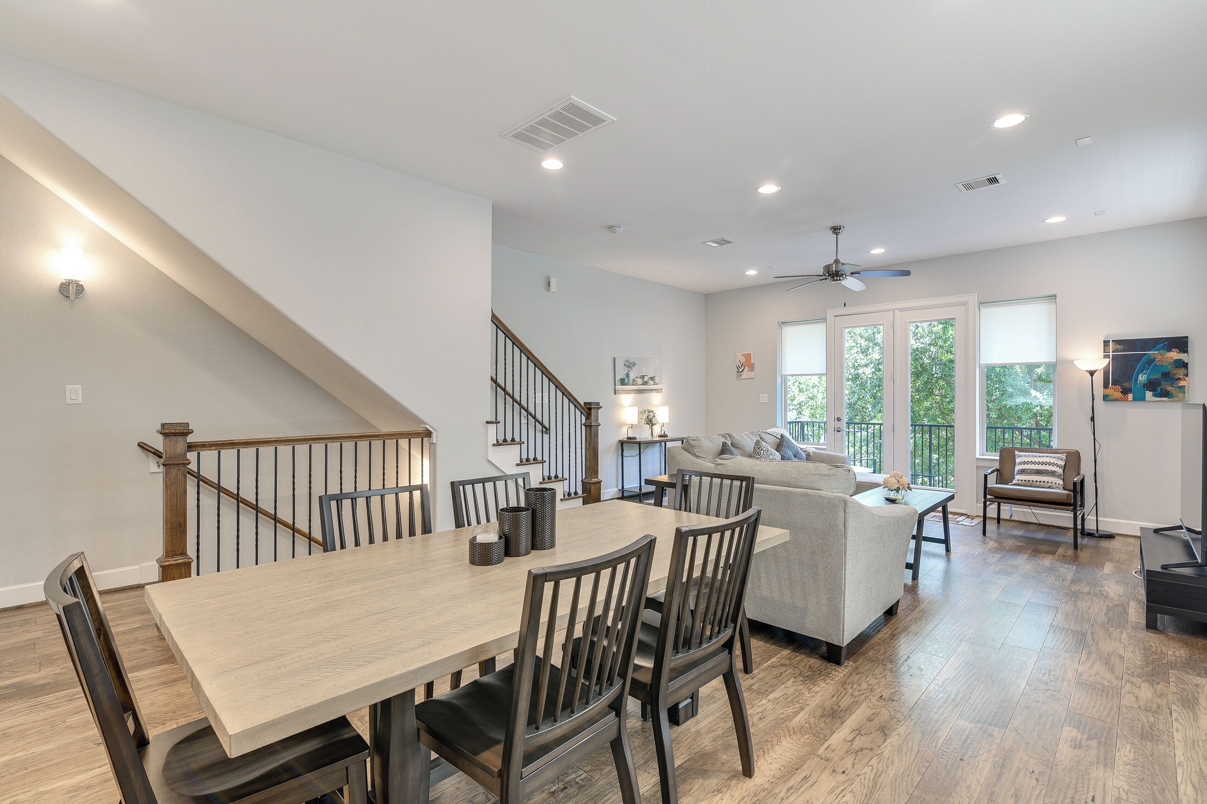 A spacious dining area with a wooden table and chairs, a staircase, a living area with a sofa and a TV, and large windows with a view of greenery.